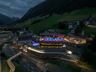 Front view of the illuminated Silvretta Therme in Ischgl at night with surrounding residential buildings.